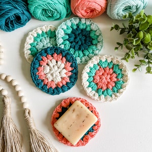 May include: Four crocheted coasters in shades of teal, pink, and white. The coasters are round and have a raised, textured design. A bar of soap is sitting on top of one of the coasters.