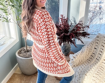 May include: A rust and cream-colored knit cardigan with a geometric pattern. The long-sleeved sweater has a cream-colored belt and pockets. The person is wearing blue jeans and brown sandals. The background includes a window with a snowy view and a large potted plant.