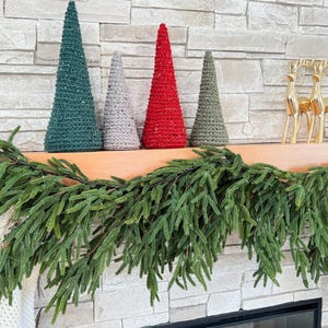 May include: Four crocheted cone-shaped Christmas trees in green, gray, red, and brown colors, arranged on a wooden mantel with a green garland draped over the front.