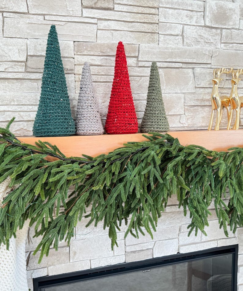 May include: Four crocheted cone-shaped Christmas trees in green, gray, red, and brown colors, arranged on a wooden mantel with a green garland draped over the front.