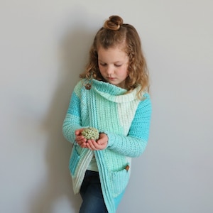 May include: A young person wearing a blue and green crocheted cardigan with a cowl neck. The cardigan has a button closure and pockets. The person is holding a small, round, green object in their hands.