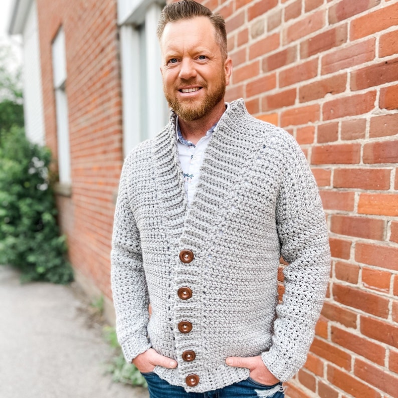 May include: A grey crocheted cardigan with a collar and button fastening. The cardigan is being worn by a person with a beard and is standing in front of a brick wall.