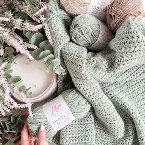Puede incluir: Una manta de ganchillo color verde salvia con un patr&oacute;n de puntos texturizados, acompa&ntilde;ada de tres ovillos de hilo MJs Off the Hook Designs Timeless Tones anti-pilling DK en verde salvia, beige y marr&oacute;n claro, con etiquetas de papel blanco.