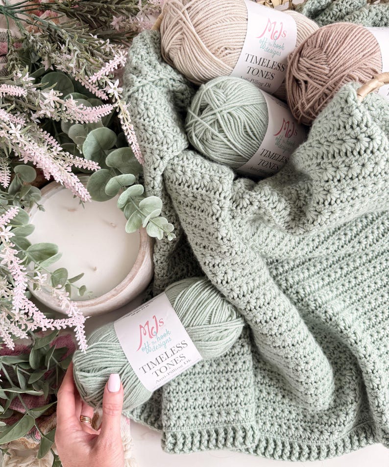 Puede incluir: Una manta de ganchillo color verde salvia con un patr&oacute;n de puntos texturizados, acompa&ntilde;ada de tres ovillos de hilo MJs Off the Hook Designs Timeless Tones anti-pilling DK en verde salvia, beige y marr&oacute;n claro, con etiquetas de papel blanco.