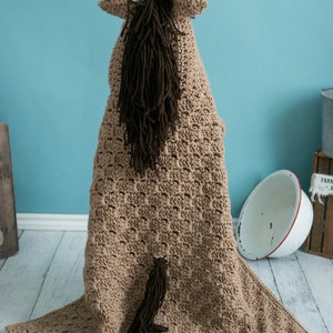 Pu&ograve; includere: Una coperta per cavallo in crochet marrone con una lunga coda e criniera marrone. La coperta &egrave; stesa su un pavimento in legno chiaro.