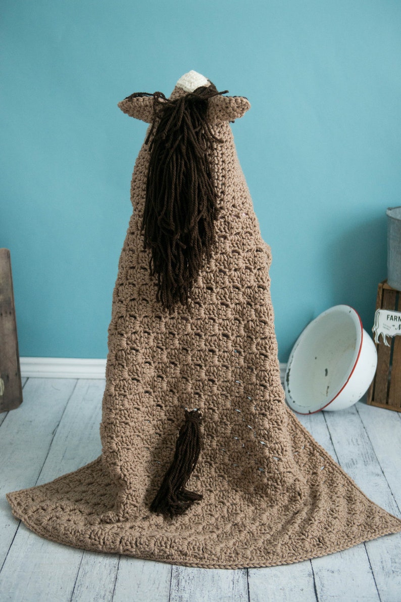 Pu&ograve; includere: Una coperta per cavallo in crochet marrone con una lunga coda e criniera marrone. La coperta &egrave; stesa su un pavimento in legno chiaro.