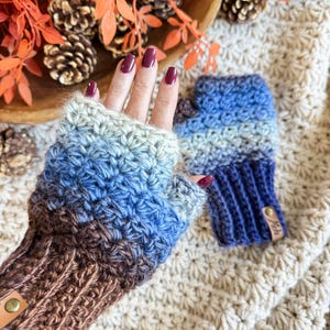 May include: Crochet fingerless gloves in blue, brown, and grey. The gloves feature a ribbed cuff and a button detail. The image shows the gloves on a cream-coloured, textured surface, with autumn-themed decorations in the background.