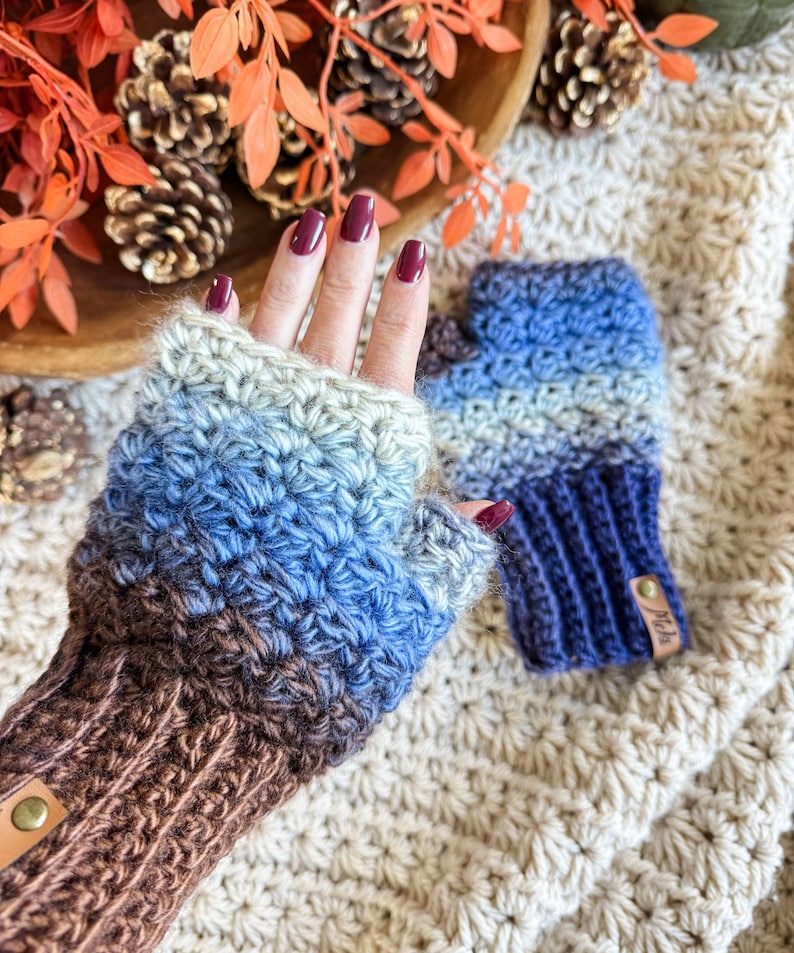 Peut inclure: Mitaines sans doigts crochet&eacute;es &agrave; la main, dans des tons de bleu, marron et gris. Les gants ont un poignet c&ocirc;tel&eacute; et un motif de point textur&eacute;. Une petite &eacute;tiquette en cuir est attach&eacute;e au poignet. Les gants sont pr&eacute;sent&eacute;s sur une surface textur&eacute;e de couleur cr&egrave;me.