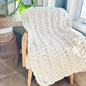 Peut inclure: Une couverture en tricot blanc &eacute;paisse drap&eacute; sur une chaise en bois. La couverture est faite de fil &eacute;pais et a une texture douce et moelleuse.