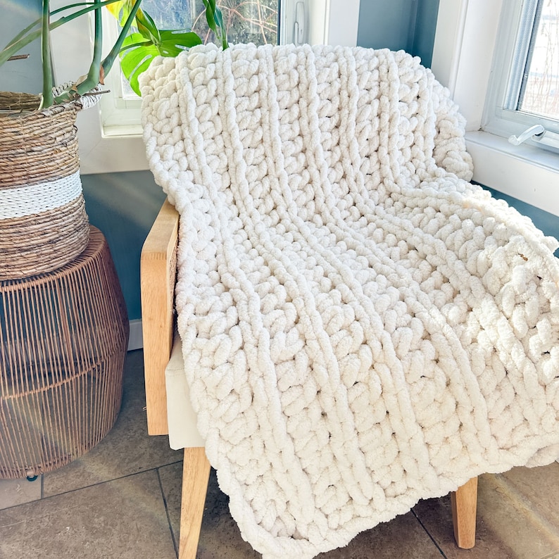 Peut inclure: Une couverture en tricot blanc &eacute;paisse drap&eacute; sur une chaise en bois. La couverture est faite de fil &eacute;pais et a une texture douce et moelleuse.