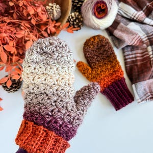 Peut inclure: Une paire de mitaines crochet&eacute;es &agrave; la main dans un d&eacute;grad&eacute; de couleurs, du cr&egrave;me au violet, avec un poignet orange. La deuxi&egrave;me mitaine est dans les tons orange et marron. Une pelote de laine et une &eacute;charpe &agrave; carreaux sont en arri&egrave;re-plan.
