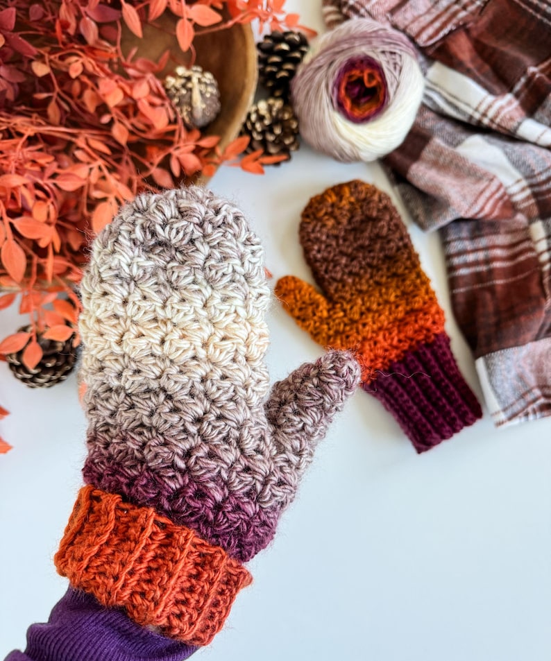 Peut inclure: Une paire de mitaines crochet&eacute;es &agrave; la main dans un d&eacute;grad&eacute; de couleurs, du cr&egrave;me au violet, avec un poignet orange. La deuxi&egrave;me mitaine est dans les tons orange et marron. Une pelote de laine et une &eacute;charpe &agrave; carreaux sont en arri&egrave;re-plan.