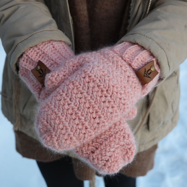 May include: A pair of pink crocheted mittens with brown leather tags that have a triangle symbol and the letter "U".