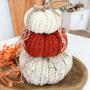 K&ouml;nnte beinhalten: Ein Stapel aus drei gestrickten K&uuml;rbissen in Creme, Orange und Beige, angeordnet auf einem Holztablett. Die K&uuml;rbisse sind aus dickem Garn gefertigt und mit Bindfaden gebunden. Ein Etikett mit der Aufschrift "Pumpkin" ist angebracht.
