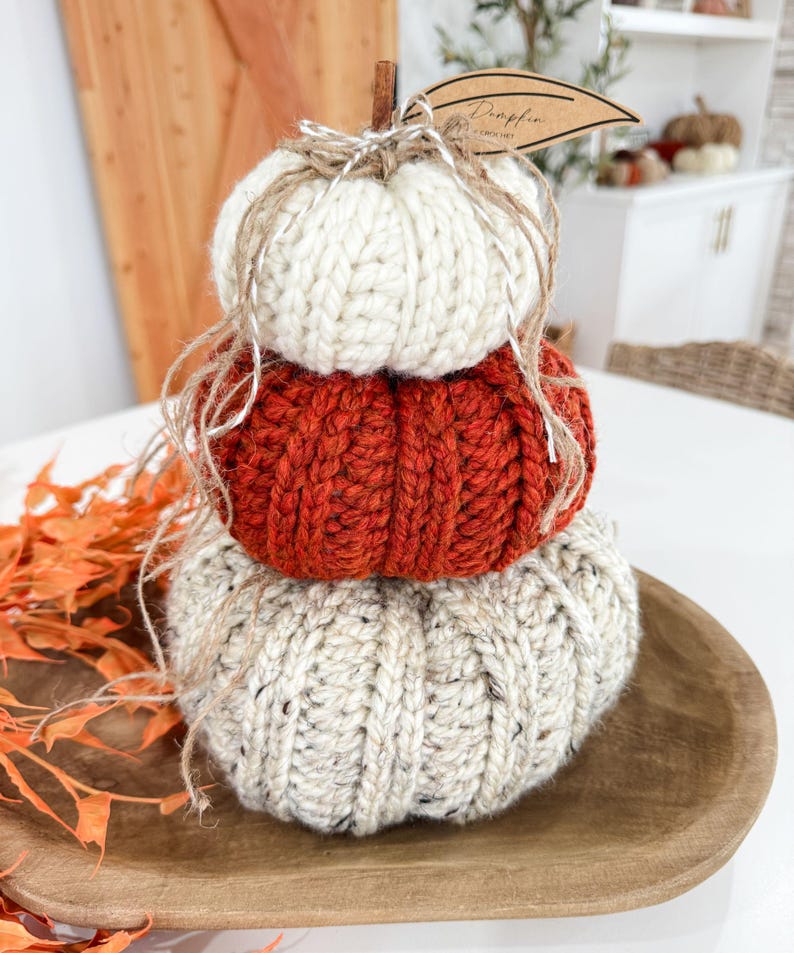 K&ouml;nnte beinhalten: Ein Stapel aus drei gestrickten K&uuml;rbissen in Creme, Orange und Beige, angeordnet auf einem Holztablett. Die K&uuml;rbisse sind aus dickem Garn gefertigt und mit Bindfaden gebunden. Ein Etikett mit der Aufschrift "Pumpkin" ist angebracht.