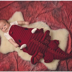 Pode incluir: Um beb&ecirc; vestindo um traje de drag&atilde;o vermelho de croch&ecirc; com detalhes brancos. O beb&ecirc; est&aacute; deitado em uma superf&iacute;cie branca e fofa.