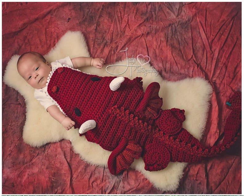Pode incluir: Um beb&ecirc; vestindo um traje de drag&atilde;o vermelho de croch&ecirc; com detalhes brancos. O beb&ecirc; est&aacute; deitado em uma superf&iacute;cie branca e fofa.