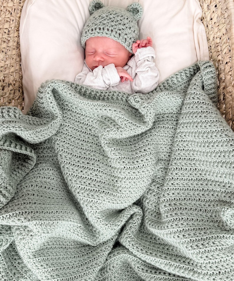 Puede incluir: Una manta de beb&eacute; tejida a ganchillo en color verde salvia con un patr&oacute;n de punto de estrella texturizado, acompa&ntilde;ada de un gorro de beb&eacute; a juego en ganchillo verde salvia con dos peque&ntilde;as orejas redondeadas en la parte superior.