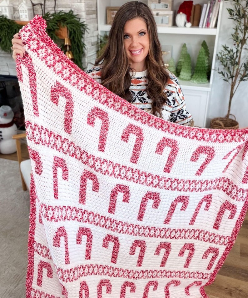K&ouml;nnte beinhalten: Eine wei&szlig;-rote H&auml;keldecke mit einem Zuckerstangenmuster. Die Decke hat einen roten Rand und Zuckerstangenmuster. Die Decke wird hochgehalten und zeigt das festliche Weihnachtsthema.