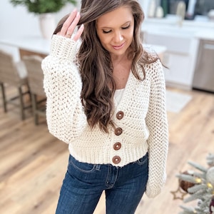 Może przedstawiać: Kremowy, gruby sweter z dekoltem w serek i czterema dużymi, okrągłymi, brązowymi guzikami. Kardigan ma długie rękawy i jest połączony z ciemnoniebieskimi dżinsami. Ogólny styl jest swobodny i wygodny.