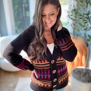 Könnte beinhalten: Schwarze Häkeljacke mit farbenfrohem geometrischem Muster in Orange-, Lila- und Rottönen. Die Strickjacke hat einen V-Ausschnitt, lange Ärmel und Holzknöpfe. Die Ärmel haben ein Streifendesign. Die Strickjacke wird über einem weißen Oberteil getragen.