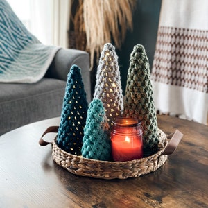 Op de afbeelding: Een set van vier gehaakte kerstboompjes in tinten groen en wit, gerangschikt in een geweven mand met een rode kaars in het midden.