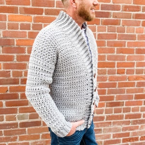 May include: A grey crocheted cardigan with a high collar and button fastening. The cardigan is being worn by a person standing in front of a brick wall.