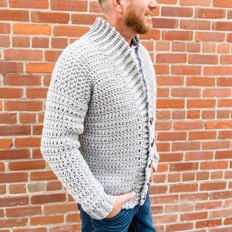 May include: A grey crocheted cardigan with a high collar and button fastening. The cardigan is being worn by a person standing in front of a brick wall.