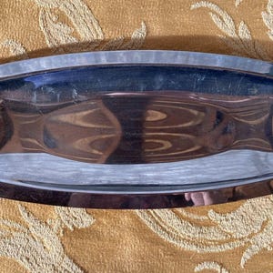 Puede incluir: Una bandeja rectangular con interior plateado pulido y asas de madera. Las asas de madera son de color marrón oscuro y están unidas a la bandeja con pequeños remaches plateados. La bandeja está sobre una superficie dorada estampada.