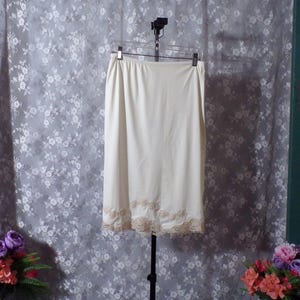 May include: A cream-colored slip skirt hangs on a hanger. The skirt has a straight cut and a delicate lace trim at the hem. The fabric appears smooth and opaque, with an elastic waistband.