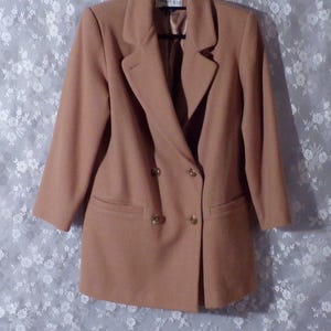 May include: A tan double-breasted blazer with gold-colored buttons and two front pockets. The blazer has a classic lapel design and is made of a smooth fabric. The blazer is hanging on a hanger.
