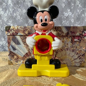 Peut inclure: Une figurine de jouet vintage de Mickey Mouse en tenue de chef. La figurine est principalement jaune, rouge, noire et blanche. Mickey tient un objet rouge et jaune. Un accessoire jaune en forme de cuillère est devant la figurine.