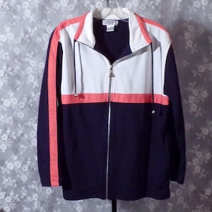 May include: A navy blue zip-up track jacket with white and coral accents. The jacket features a white panel across the chest and coral stripes on the sleeves and chest. The jacket is hanging on a hanger.