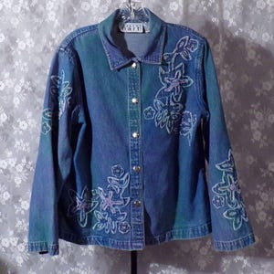 May include: A blue denim jacket with floral embroidery. The embroidery is in shades of pink, purple, and green. The jacket has a button-up front and long sleeves.