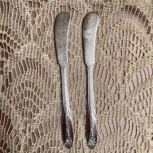 Può includere: Due coltelli da burro d'argento con manici decorati, su una superficie di uncinetto beige.