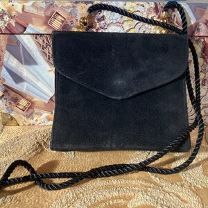 Könnte beinhalten: Eine schwarze Wildleder-Umhängetasche mit Klappenverschluss. Die Tasche hat einen schwarzen Seilriemen mit goldfarbenen Akzenten. Die quadratische Tasche scheint vintage zu sein. Der Hintergrund umfasst eine dekorative Box und eine gemusterte Oberfläche.
