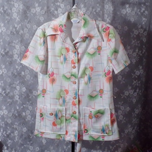 May include: White short-sleeved shirt with a colorful geometric pattern. The shirt features a collared neckline, button-down front, and two front pockets. The pattern includes diamonds, circles, and lines in shades of orange, green, pink, and blue.
