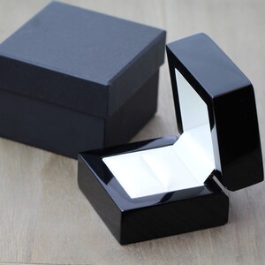 May include: A black ring box with a white interior and a hinged lid. The box is open and the lid is propped up.