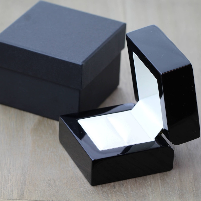 May include: A black ring box with a white interior and a hinged lid. The box is open and the lid is propped up.