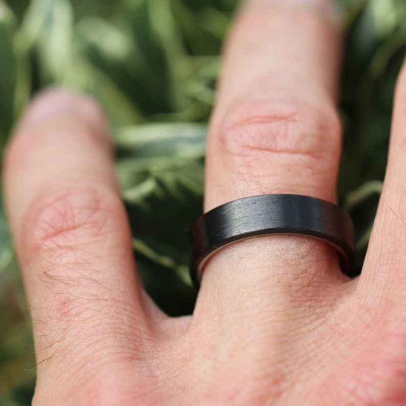 May include: A black ring with a brushed finish. The ring is worn on a finger.