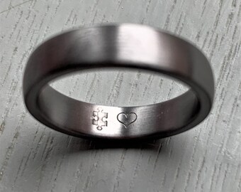 Op de afbeelding: Een grijze metalen ring met een afgeronde band. De ring heeft een puzzelstukje en een hartje aan de binnenkant gegraveerd. Geborstelde afwerking.