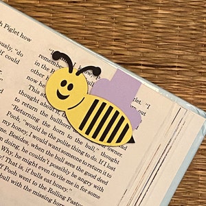 May include: A yellow and black paper bee bookmark with a smiling face. The bookmark is placed on a page of a book with the text "Returning the horn to the bull..."