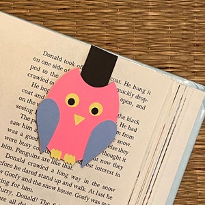 May include: A pink and blue paper owl bookmark with yellow eyes. The bookmark is on a page of a book with text about a penguin.