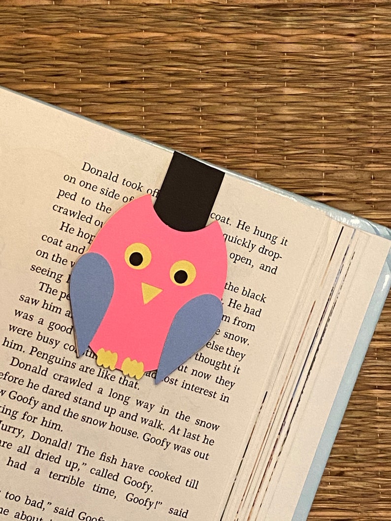 May include: A pink and blue paper owl bookmark with yellow eyes. The bookmark is on a page of a book with text about a penguin.