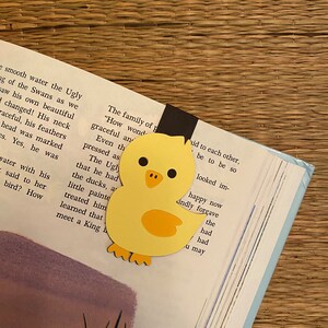 May include: A yellow paper cutout of a chick with black eyes and a black beak. The chick is holding a black bookmark with the text "The family of p" and "How wond" visible. The chick is on top of a book with text about a swan.