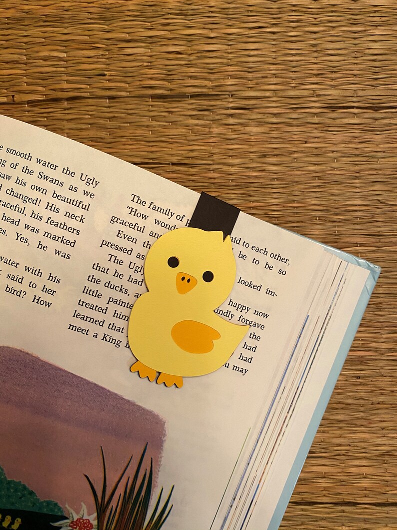 May include: A yellow paper cutout of a chick with black eyes and a black beak. The chick is holding a black bookmark with the text "The family of p" and "How wond" visible. The chick is on top of a book with text about a swan.