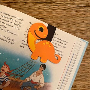 May include: Orange dinosaur-shaped bookmark with a black background. The bookmark is on top of an open book with text that reads "We the wa shi" and "look."