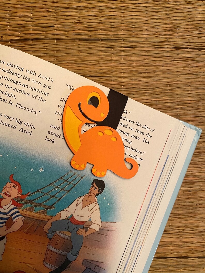 May include: Orange dinosaur-shaped bookmark with a black background. The bookmark is on top of an open book with text that reads "We the wa shi" and "look."
