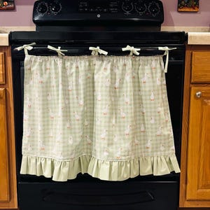 May include: A light green and white gingham curtain featuring a repeating pattern of small white geese. The curtain is attached to the oven handle with five fabric ties and features a solid light green ruffled hem along the bottom edge.