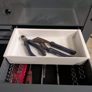 May include: A partially open gray tool chest drawer with a white tray holding pliers. Below, another drawer reveals a collection of tools, including sockets and screwdrivers. The tools are neatly organized, suggesting a workshop or garage setting.